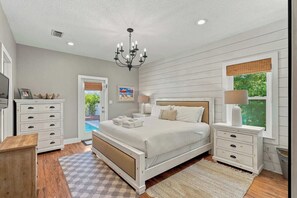 A bedroom with a bed, bedside tables, a chandelier, a window with blinds, and a view of the outdoors. - Beach cottage w/ heated pool & fenced yard (Panama City Beach)