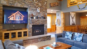 A cozy living room with a stone fireplace, a flat-screen TV, and a comfortable seating area. - PNW Luxe Lodge- 4 Suites, Game Rm, Loft, 12xSHARC, Bikes, Hot Tub, Big Deck, ADA (Sunriver)