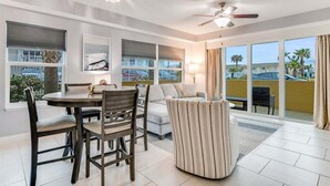 A modern hotel room with a dining area, a sofa, and a balcony with a view of palm trees and a pool.