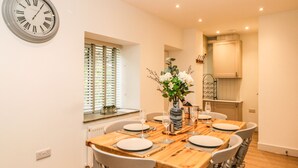 Comedor con una mesa de madera dispuesta para seis personas, un reloj en la pared y una ventana con persianas.