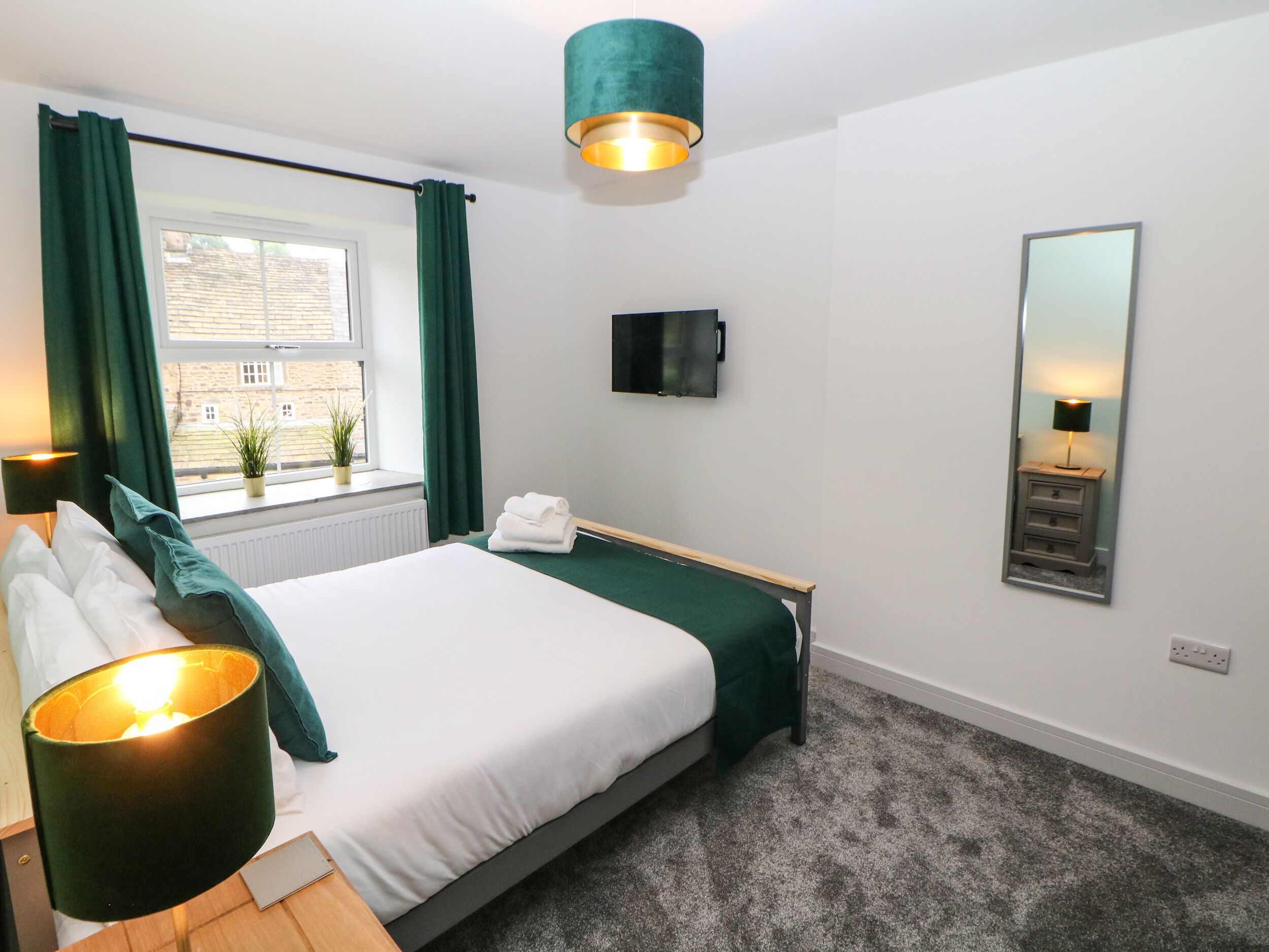 A hotel room with a bed, a bedside table with a lamp, a window with curtains, a wall-mounted TV, and a mirror.