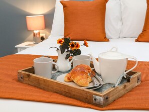 A breakfast tray with a croissant, two mugs, a teapot, and a pitcher with flowers on a bed with orange pillows and a nightstand with a lamp. - 1 Orchard View (Hope Valley)