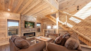 A cozy cabin interior with wooden walls, a stone fireplace, a TV, and a staircase. - New Luxury 5 BR Cabin w/ Incredible Views, Hot Tub, Wi-Fi, Arcade, Pool Table (Sevierville)