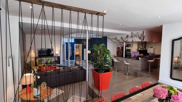 A modern living room with a sofa, a coffee table, and a large potted plant. There is a kitchen area with a counter and a sink.