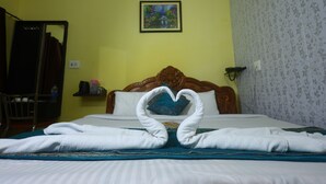 Una habitación de hotel con una cama, una toalla dispuesta en forma de corazón, un cabecero de madera, un cuadro en la pared y un papel tapiz estampado con motivos florales.