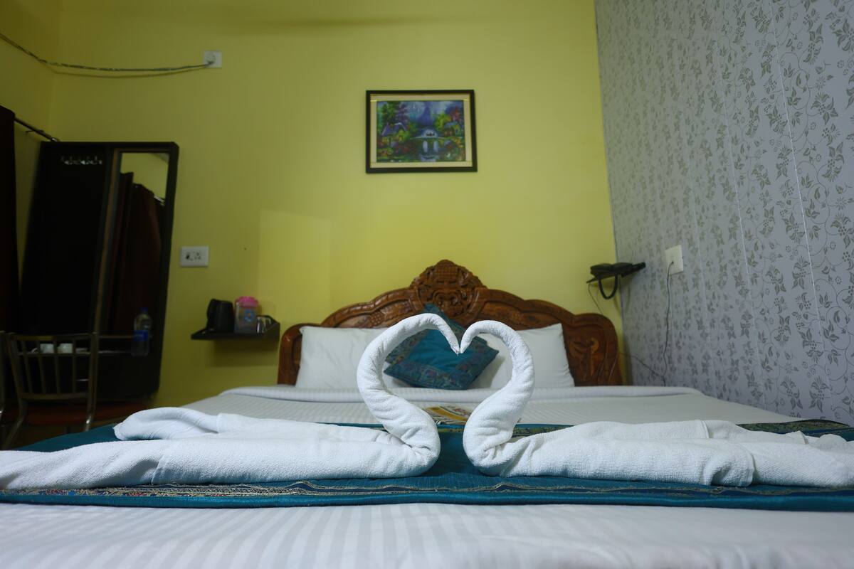 Une chambre d’hôtel avec un lit, une disposition de serviettes en forme de cœur, une tête de lit en bois, un tableau au mur et un papier peint à motifs floraux.