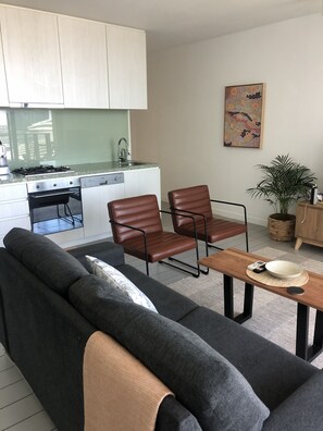 A modern kitchen with white cabinets, a built-in oven, and a stainless steel range hood. There is a dining area with a wooden table and two brown leather chairs. A gray sofa and a small rug are also visible. - Harbour views at Docklands, Melbourne. Next to Marvel stadium (Docklands)
