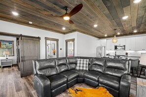 A modern living room with a leather sectional sofa, a wooden ceiling, and a kitchen in the background.