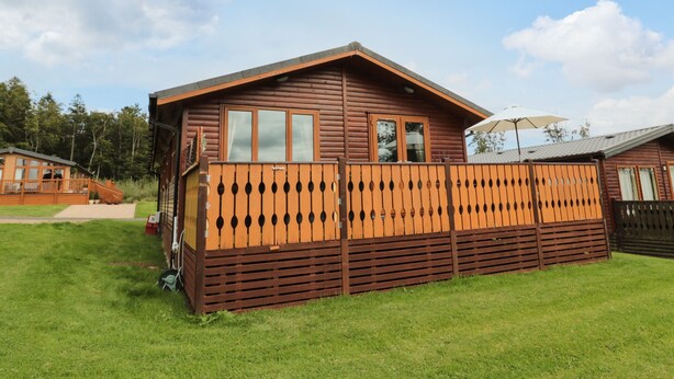 A wooden cabin with a fenced deck, a staircase leading to the entrance, and a clear sky in the background. - Hillside View (Morpeth)