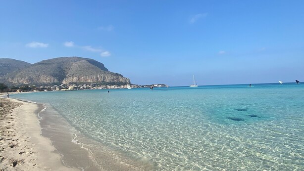 A beach with clear shallow waters, a sandy shore, and distant mountains.