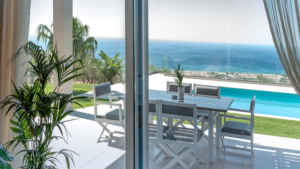 A glass-enclosed patio with a table and chairs, overlooking a pool and ocean view. - Villa Samiram (Ierapetra)