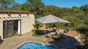 A pool area with lounge chairs and an umbrella, surrounded by a landscaped garden and a building with a tiled patio.