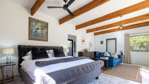 A bedroom with a large bed, a ceiling fan, wooden beams, a desk, a chair, a sofa, and a window with a view.