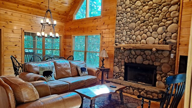 A cozy log cabin interior with a leather sofa, a stone fireplace, and a large window.