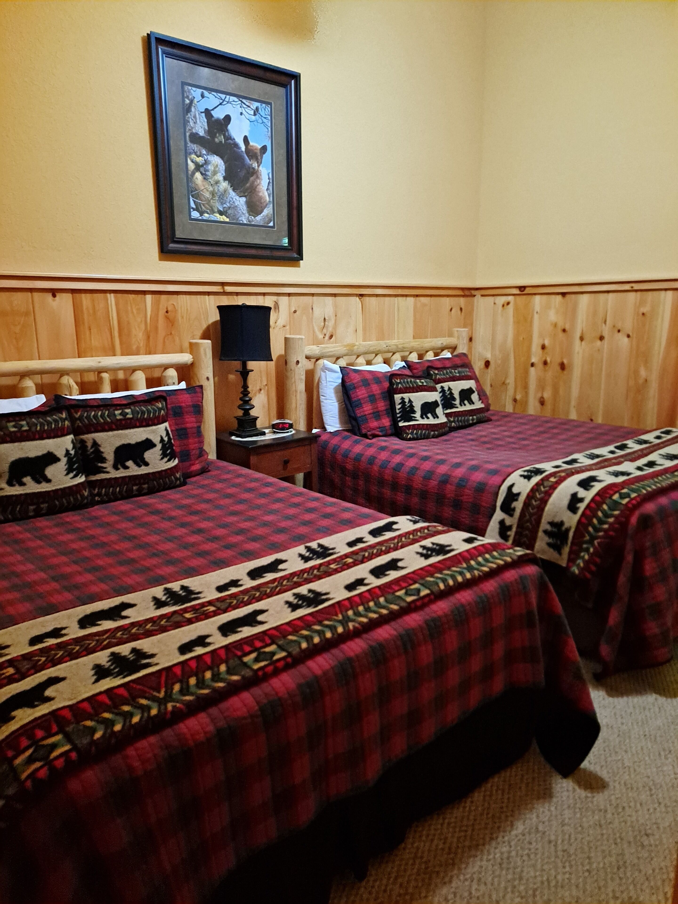 A room with two beds, each with a red and black plaid bedspread featuring bear and tree patterns. There is a framed picture of bears on the wall above the beds.