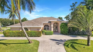 A single-story house with a tiled roof, a front entrance with a glass door, and a well-maintained lawn with palm trees.