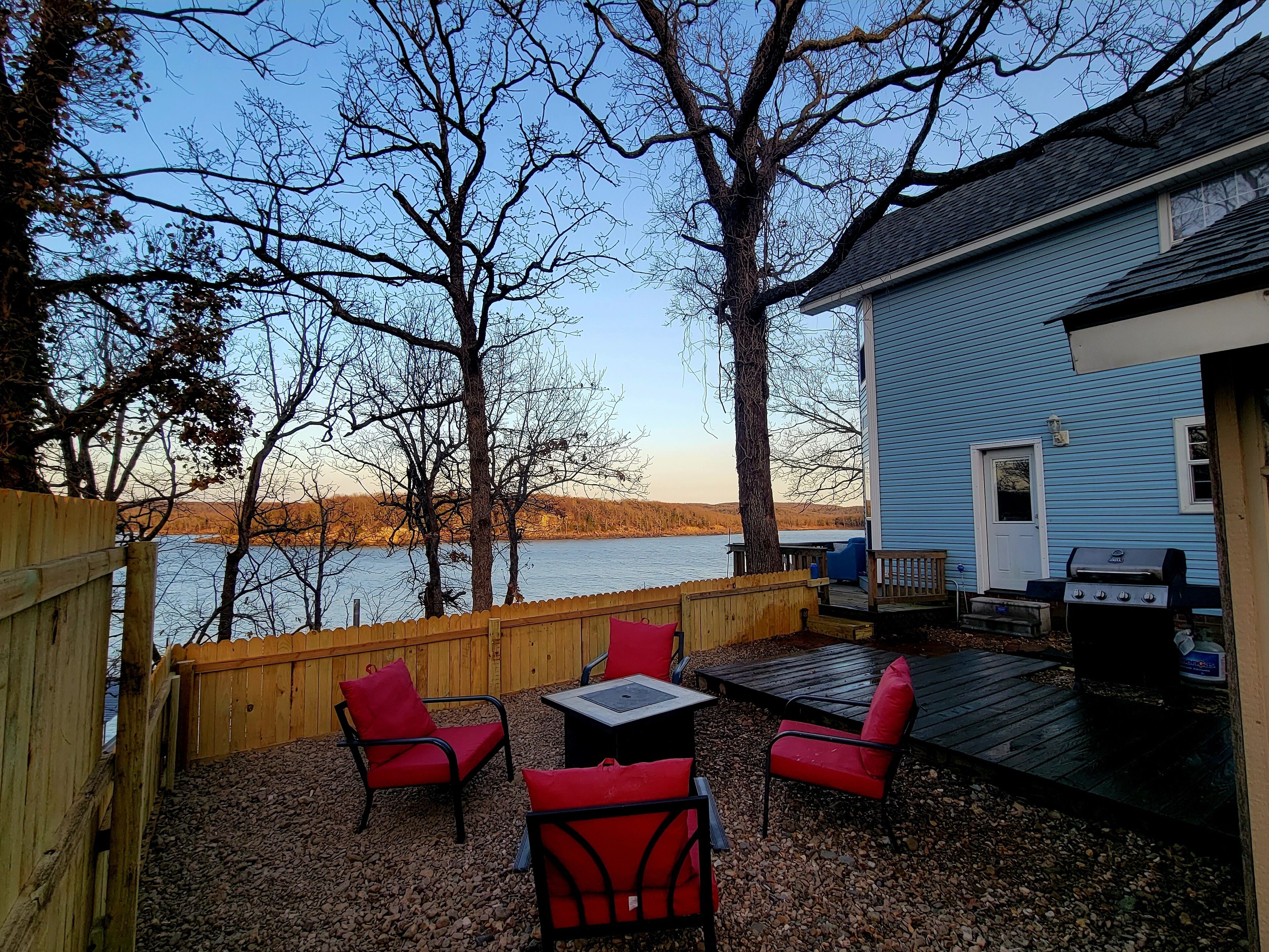 A deck with red chairs and a table, overlooking a body of water and a house with a blue exterior.