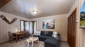 Een gezellige woonkamer met een donkergrijze bank, een klein rond tafeltje en een schilderij van een landschap. Er staat een houten eettafel met stoelen, en een raam met gordijnen.