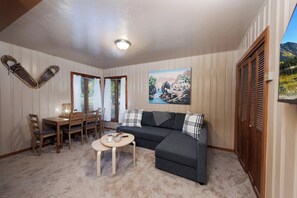 A cozy living room with a dark grey sofa, a small round table, and a painting of a landscape. There is a wooden dining table with chairs, and a window with curtains. - Pet Friendly Cabin on 3.5 acres between Durango and Purgatory - Fire Pit (Durango)