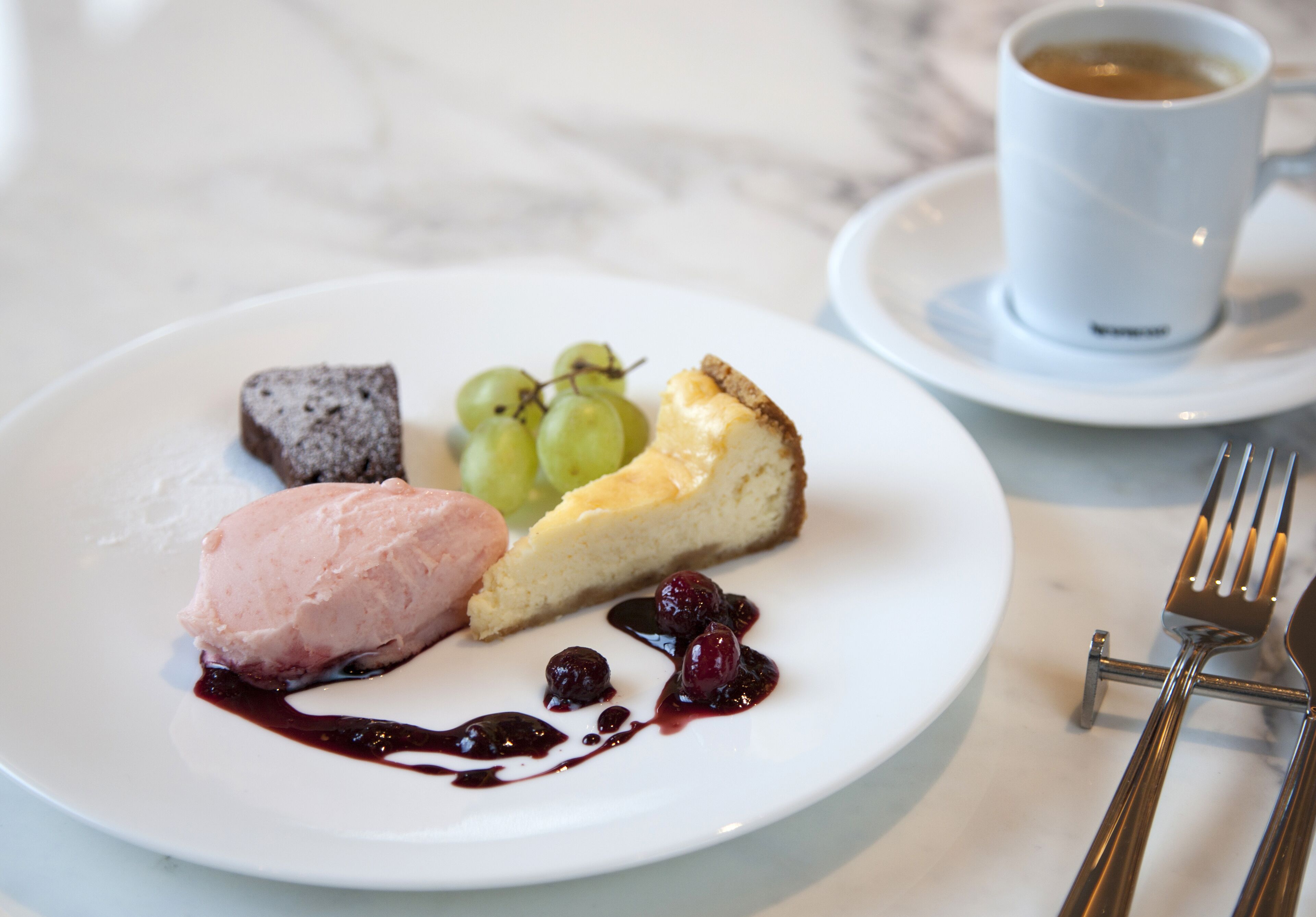 A dessert plate with ice cream, a slice of cheesecake, and grapes, accompanied by a cup of coffee on a white plate.