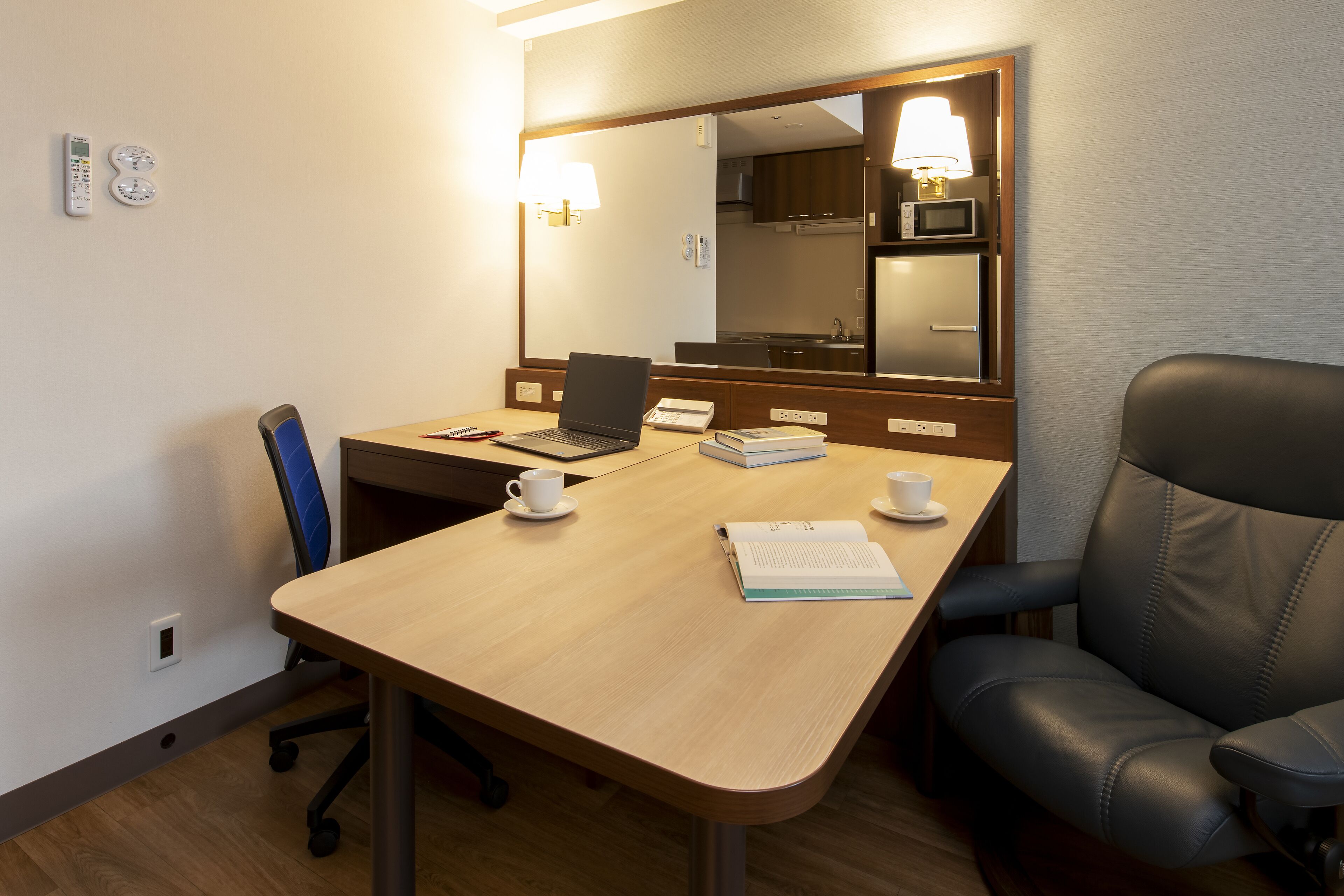 A hotel room with a wooden desk, a laptop, a chair, a microwave, and a large mirror.