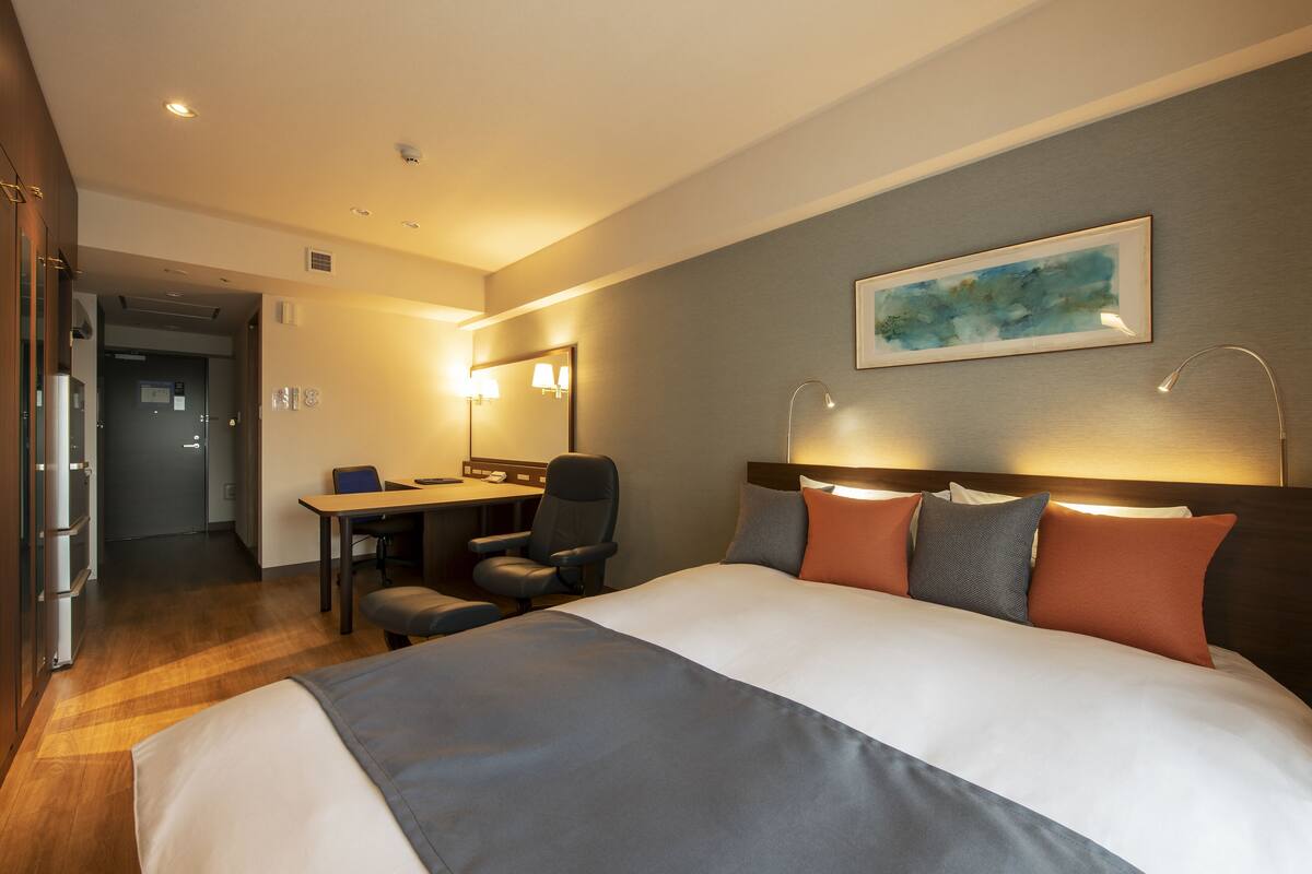 Habitació d'hotel amb un llit gran, una taula de nit i una cadira. Hi ha un mirall sobre la taula i un quadre a la paret.