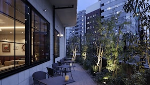 Un patio al aire libre con mesas y sillas, un pequeño jardín y vistas de la ciudad al fondo.