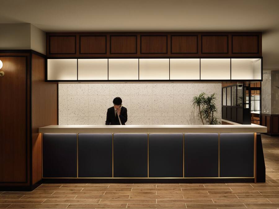 A reception area with a wooden counter, a person standing behind it, and a tiled wall behind.