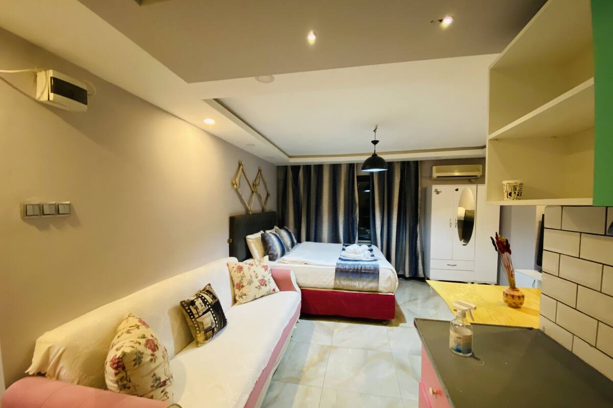 A modern hotel room with a large bed, a sofa, a kitchenette, and a bathroom.