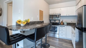 A modern kitchen with white cabinets, a stainless steel refrigerator, an oven, and a microwave. There are two black bar stools and a vase with sunflowers on the counter.