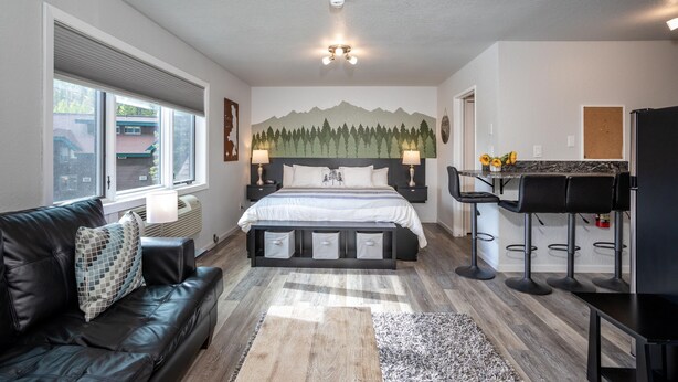 A modern bedroom with a bed, a black leather sofa, a dining area with bar stools, and a mountain mural on the wall.
