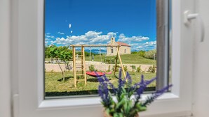 Blick aus dem Fenster auf eine Wiese mit einer Holz-Schaukelanlage und einer roten Schaukel, ein kleines Gebäude mit rotem Dach und einem strahlend blauen Himmel.