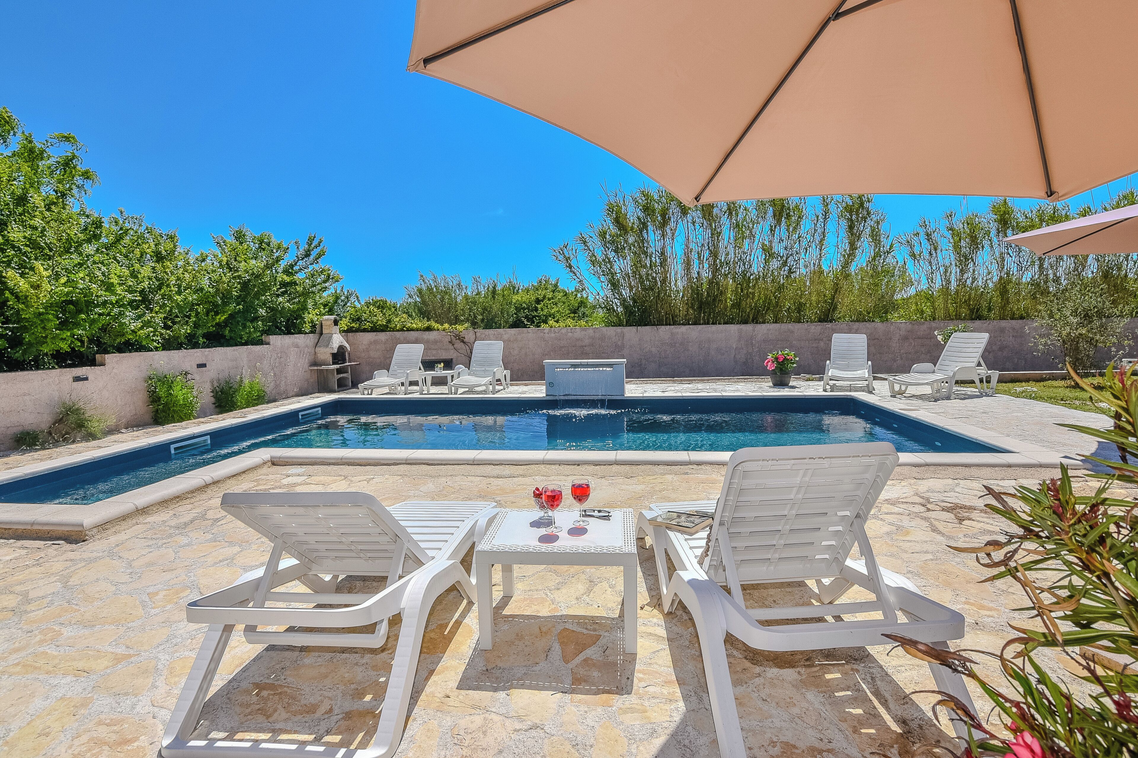 A swimming pool with lounge chairs and umbrellas, surrounded by a stone patio and greenery.