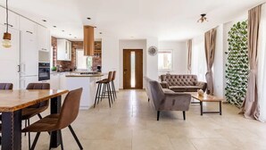Eine moderne Küche mit weißen Schränken, einer Ziegelrückwand und einer zentralen Kochinsel. Es gibt einen Essbereich mit Holztisch und -stühlen sowie ein Wohnzimmer mit grauem Sofa und Couchtisch.
