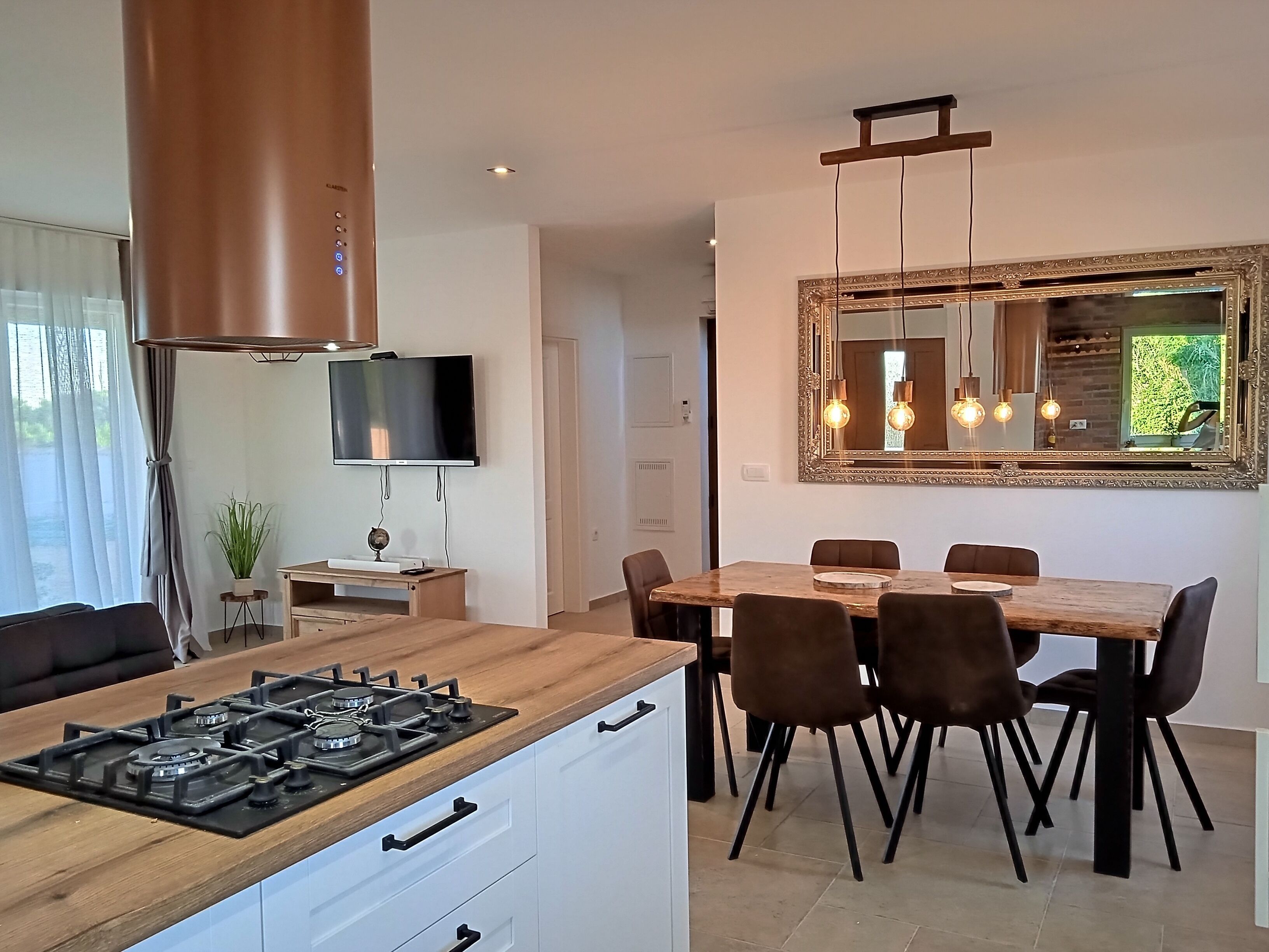 A modern kitchen with a gas stove, wooden countertop, and white cabinets. There is a dining area with a wooden table and chairs, and a large mirror above the table.