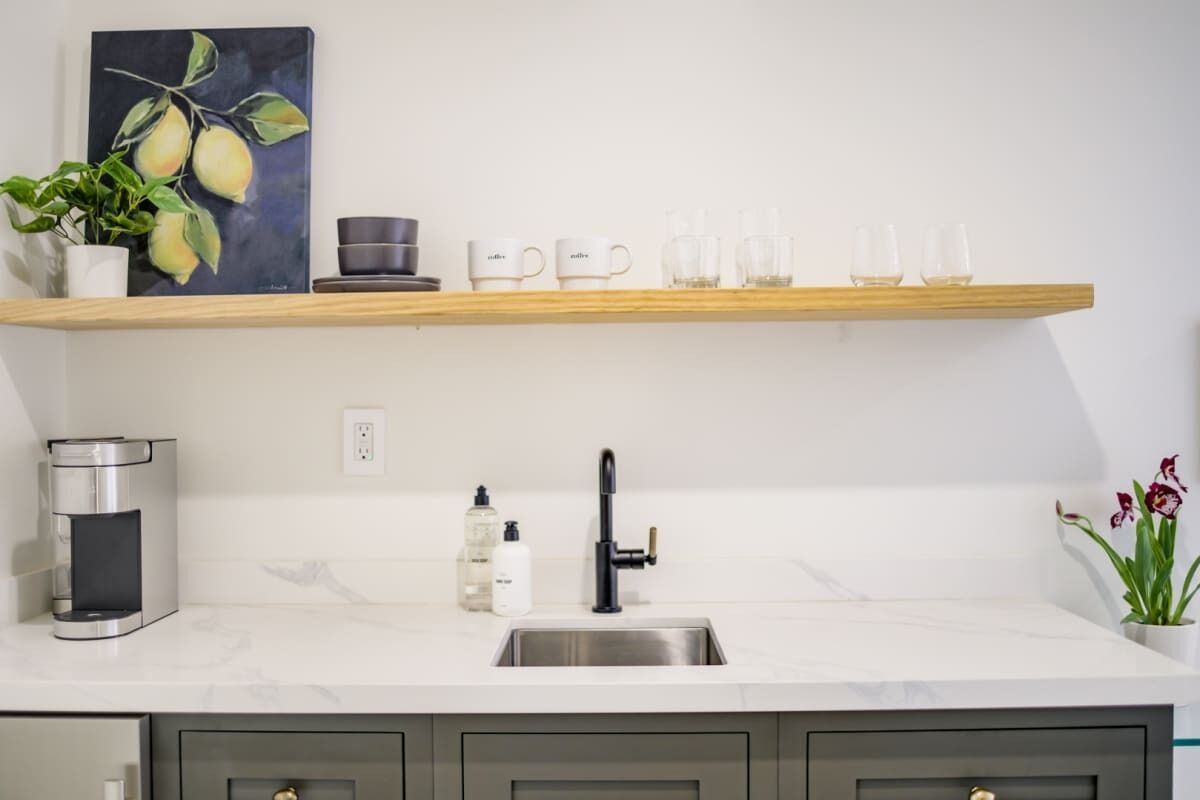 A modern kitchen with a marble countertop, a stainless steel sink, a coffee machine, a shelf with cups and glasses, and a painting of pears.