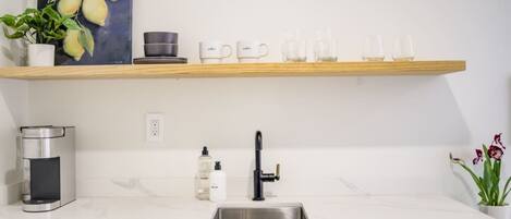 A modern kitchen with a marble countertop, a stainless steel sink, a coffee machine, a shelf with cups and glasses, and a painting of pears.
