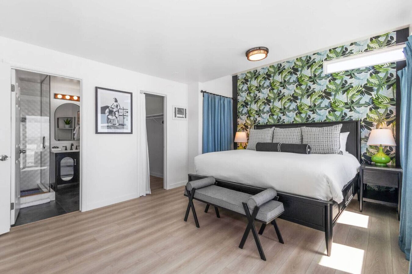 A hotel room with a bed, a bench, a mirror, a vanity, and a shower.