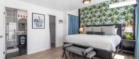 A hotel room with a bed, a bench, a mirror, a vanity, and a shower.