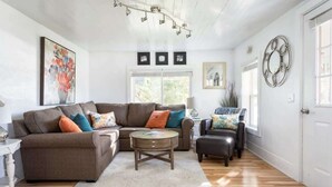 A living room with a brown sectional sofa, a round coffee table, and a white rug. There are three framed pictures on the wall above the sofa. - Ames Cottage in Elk Rapids - Dog Friendly (Elk Rapids)
