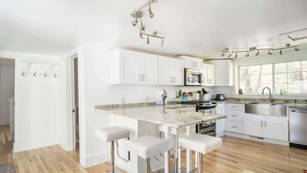 A modern kitchen with white cabinets, a granite countertop, and stainless steel appliances. There are three bar stools and a window providing natural light. - Ames Cottage in Elk Rapids - Dog Friendly (Elk Rapids)