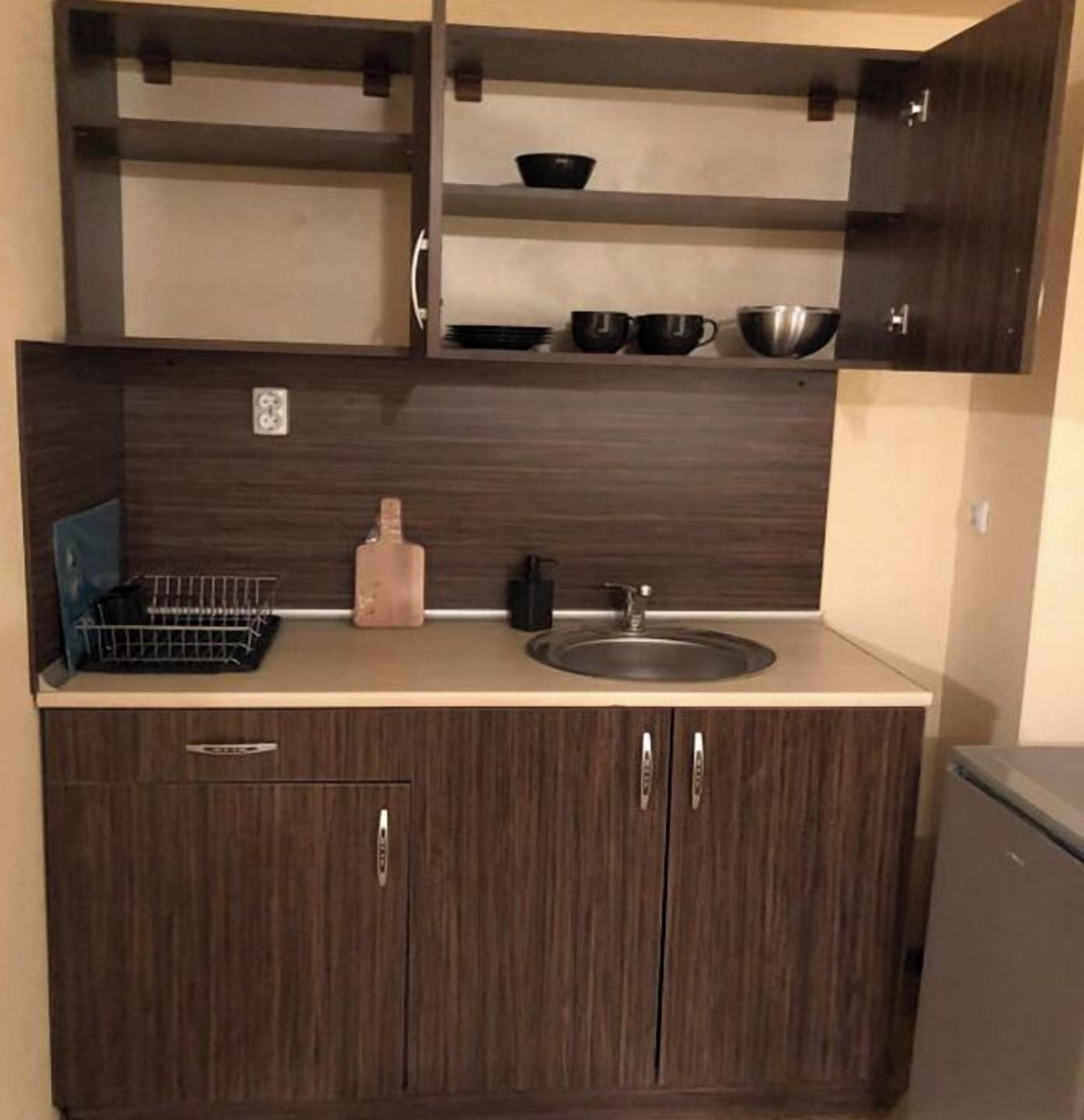 A compact kitchen with dark wood cabinets, a sink, and a countertop with a dish rack and a bottle.