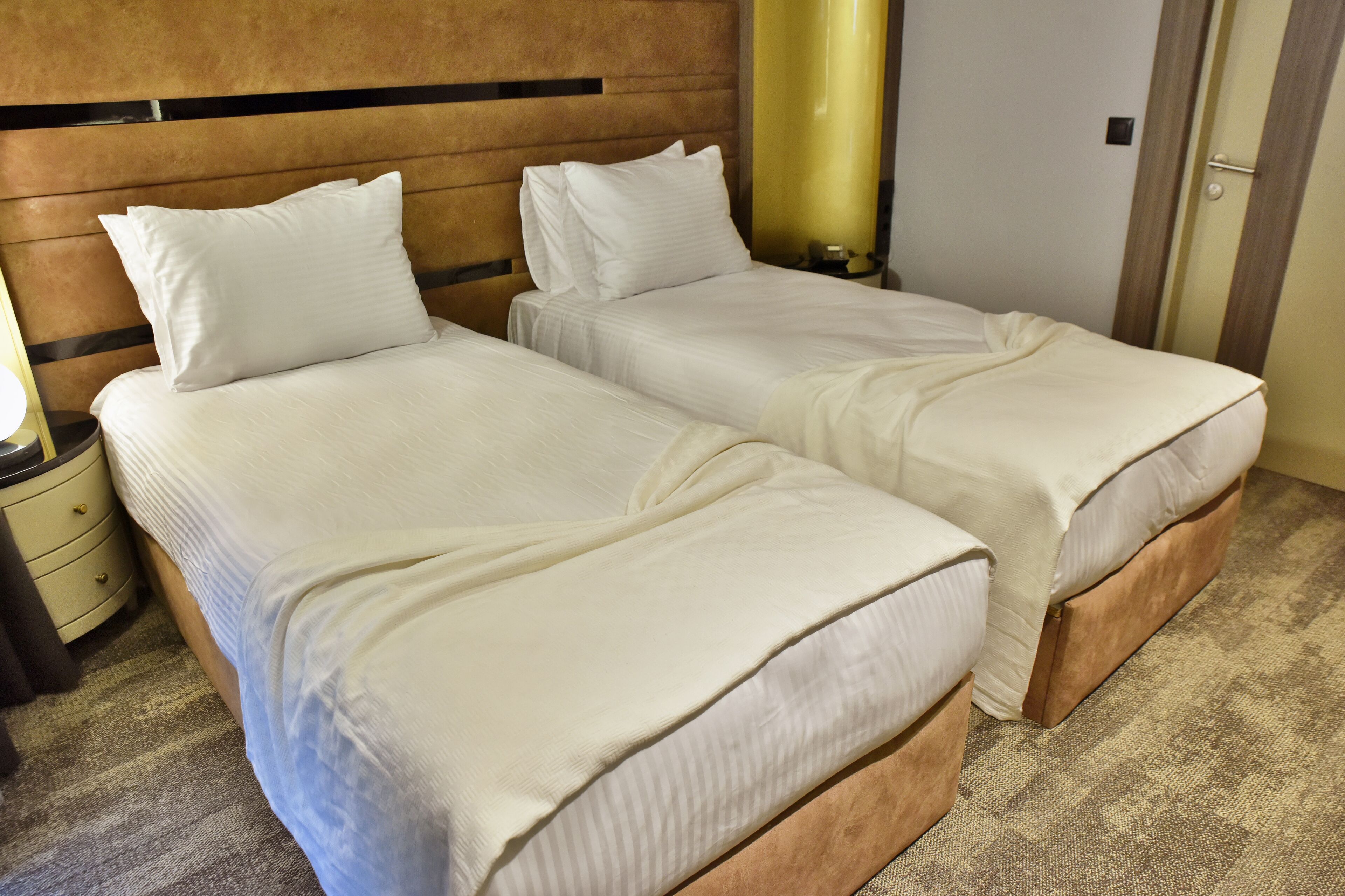 A hotel room with two beds, a wooden headboard, a nightstand with a lamp, and a carpeted floor.
