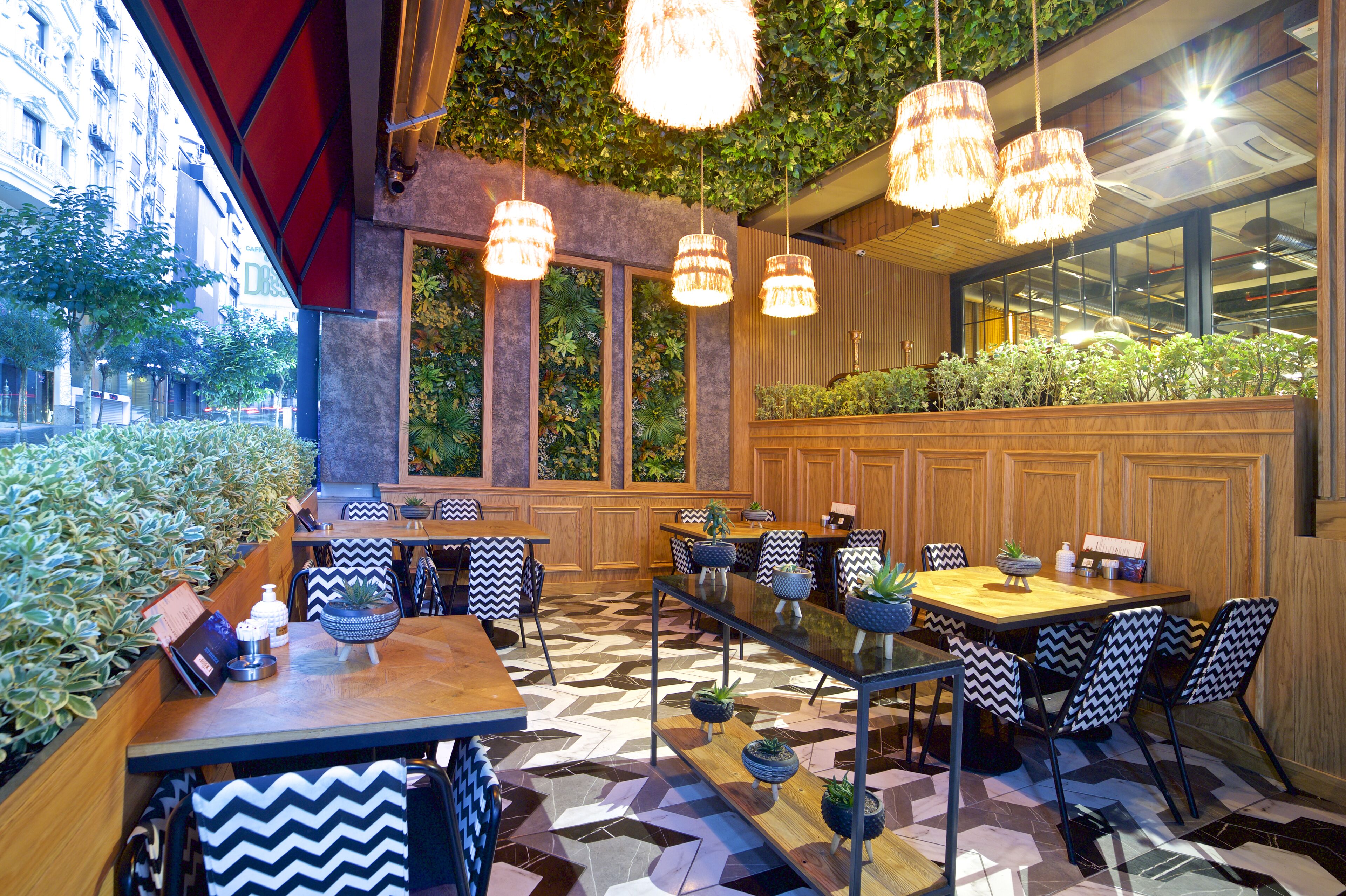 A restaurant with a modern interior, featuring a mix of wooden and glass elements, multiple dining tables with black and white patterned chairs, and hanging pendant lights.