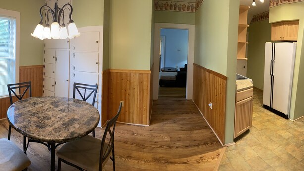 A dining area with a round table and chairs, wooden paneling, a window with blinds, a light fixture, and a kitchen with a refrigerator. - 1st Class Rentals Munson Manor VIII at 1 Columbia Street a 1 Bedroom Apartment (Oneonta)