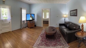 A living room with a wooden floor, a black leather couch, a round coffee table, a TV on a wooden stand, and a lamp on a side table. - 1st Class Rentals Munson Manor VIII at 1 Columbia Street a 1 Bedroom Apartment (Oneonta)
