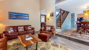 A spacious living room with a brown leather sofa, a wooden coffee table, and a large painting of a lake. A staircase leads to a kitchen area with wooden cabinets and a dining table. - DESC08 - Lovely Home, Foosball, Hot Tub, Sauna (Sunriver)