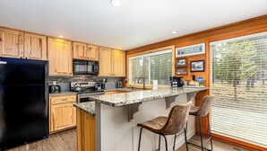 A kitchen with wooden cabinets, a black refrigerator, a microwave, and a granite countertop. There are two bar stools and a window with blinds. - DESC08 - Lovely Home, Foosball, Hot Tub, Sauna (Sunriver)