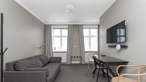 Una habitación moderna con un sofá gris, una mesa de comedor con sillas, un televisor montado en la pared y ventanas grandes con cortinas.