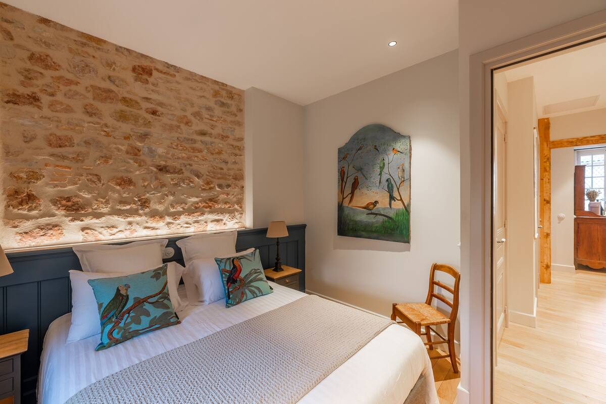 Une chambre avec un mur en pierre, un tableau représentant des oiseaux, un lit avec des oreillers et une chaise en bois.